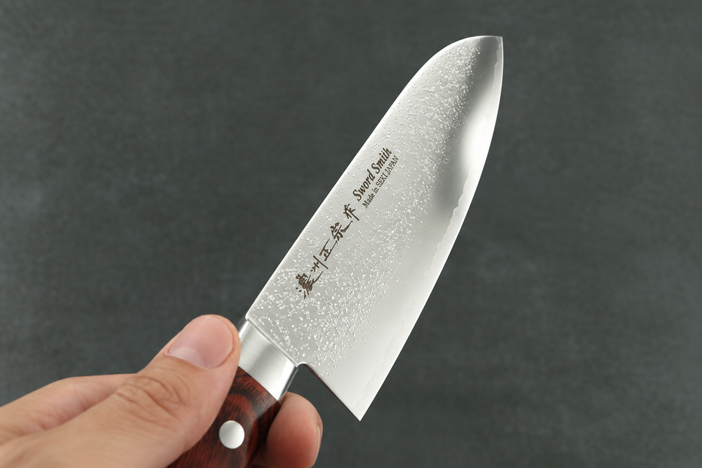 Nahaufnahme eines japanischen Santokumessers von Sword Smith mit Nashiji-Oberfläche und Kanji-Gravur, gehalten in der Hand vor dunklem Hintergrund.